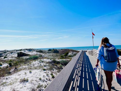 A person walks along a wooden boardwalk towards the beach, with sand dunes and the ocean in the background under a clear sky.