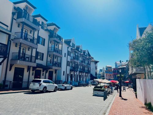 A sunny street lined with white buildings, balconies, and outdoor seating, with cars parked along the cobblestone path.
