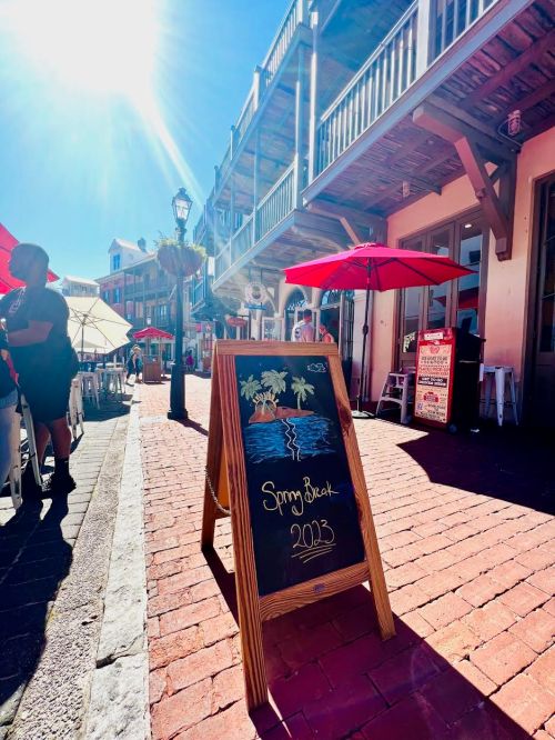 A sunny street scene with a chalkboard sign for "Spring Break 2023" and outdoor dining under umbrellas.