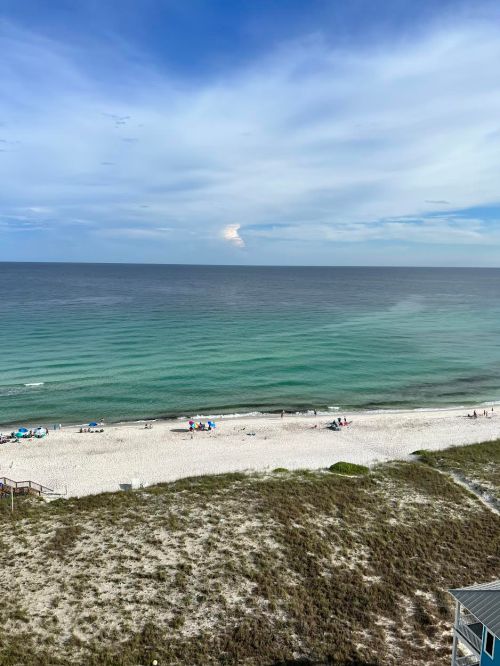 A serene beach scene with gentle waves, white sand, and a clear blue sky, dotted with a few beachgoers and umbrellas.