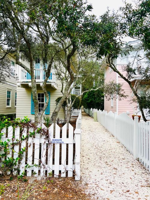 A narrow path lined with trees and a white picket fence leads to colorful houses in a serene neighborhood.