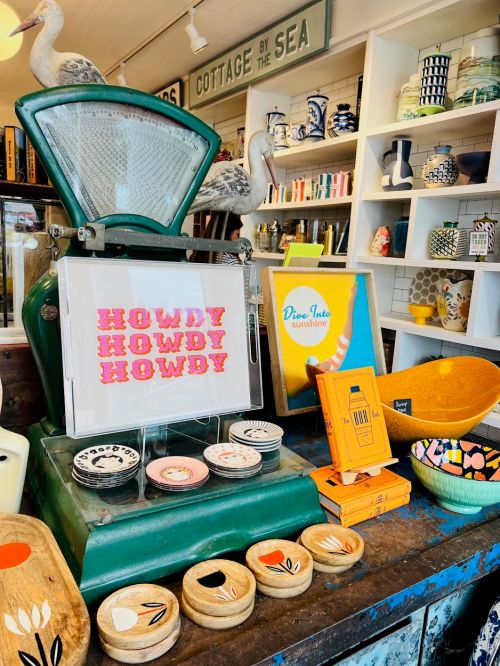 A vintage green scale displays colorful items, including bowls and decor, in a charming coastal shop setting.