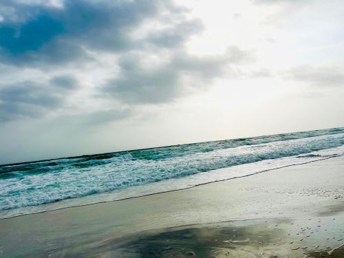 Waves gently crash on a sandy beach under a cloudy sky, reflecting light on the wet surface.
