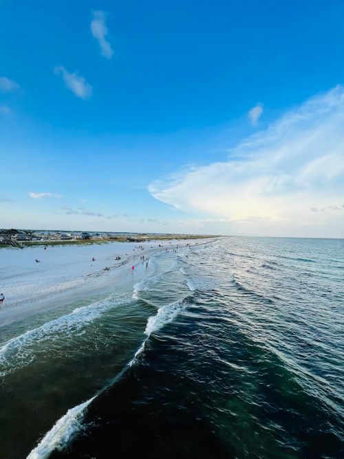 A serene beach scene with gentle waves, people walking along the shore, and a clear blue sky.