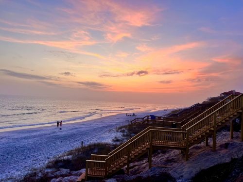 A serene beach at sunset, featuring wooden stairs leading down to the shore and silhouettes of people walking along the water.