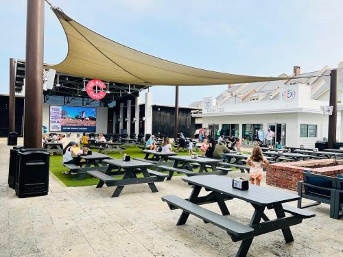 Outdoor dining area with picnic tables, a large screen, and people enjoying their meals under a shaded canopy.