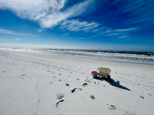 A beach scene with a striped chair on white sand, gentle waves, and a clear blue sky with wispy clouds.