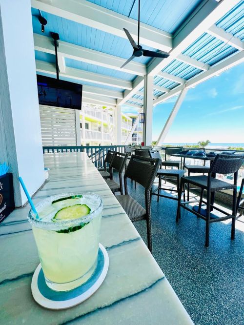 A refreshing cocktail with cucumber sits on a marble table, overlooking a sunny outdoor seating area by the water.