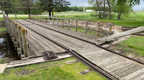A weathered wooden bridge over a small waterway, surrounded by greenery and fields in the background.
