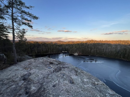 A rocky outcrop overlooks a frozen lake surrounded by trees and distant mountains under a clear sky at sunset.