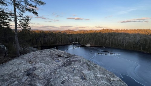 A serene landscape featuring a frozen lake surrounded by trees and mountains under a clear sky at sunset.