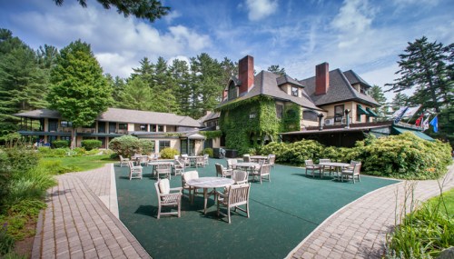 A scenic view of a lodge surrounded by greenery, featuring outdoor seating and a blue sky with clouds.