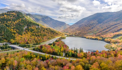 A scenic view of a lake surrounded by vibrant autumn foliage and mountains under a partly cloudy sky.