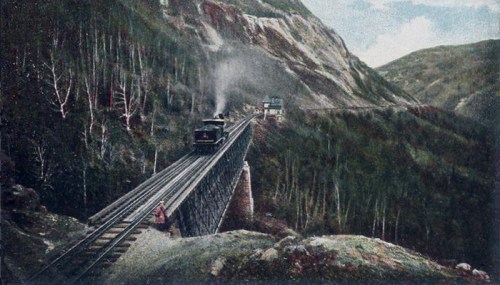 A vintage steam train travels along a bridge in a mountainous landscape, surrounded by trees and rocky cliffs.