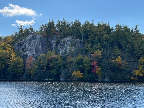 A serene lake reflects a rocky shoreline adorned with vibrant autumn foliage and a clear blue sky.