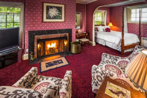 Cozy room with a fireplace, patterned chairs, a bed, and warm lighting, featuring rich red wallpaper and a rug.