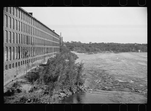 Black and white photo of a brick factory building alongside a river, with trees and rocky shoreline visible.