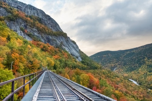 A scenic view of train tracks leading through vibrant autumn foliage and mountains under a cloudy sky.