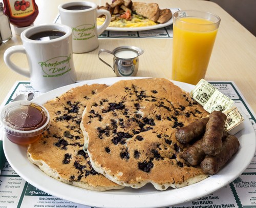 A plate of blueberry pancakes with sausage links, syrup, coffee, and orange juice on a diner table.