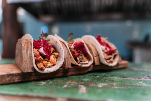 Three tacos filled with colorful toppings, served on a wooden tray against a rustic background.