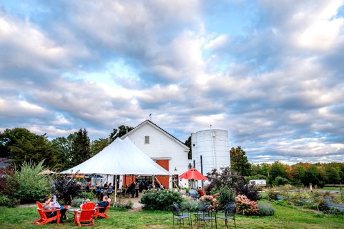 A scenic outdoor venue with a white barn, tent, and seating, surrounded by greenery and a cloudy sky.