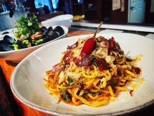 A plate of pasta topped with herbs, cheese, and a chili pepper, served alongside a bowl of mussels and greens.
