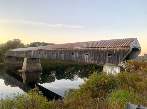 A long wooden covered bridge spans a calm river, reflecting the surrounding trees and sky at sunset.