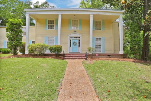 A large yellow house with blue shutters, white columns, and a front walkway surrounded by green grass and trees.