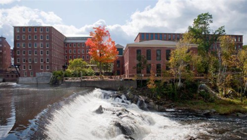 A brick building by a river with a waterfall, surrounded by trees, including one with bright red leaves.