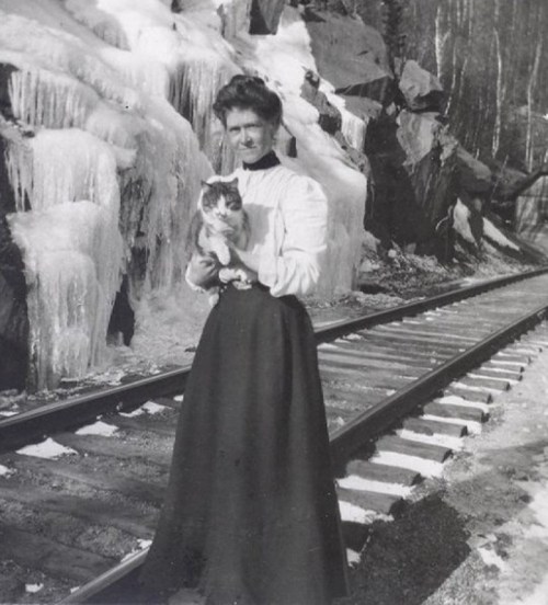 A woman in a long skirt holds a cat near icy rocks by a railway track in a vintage black-and-white photo.