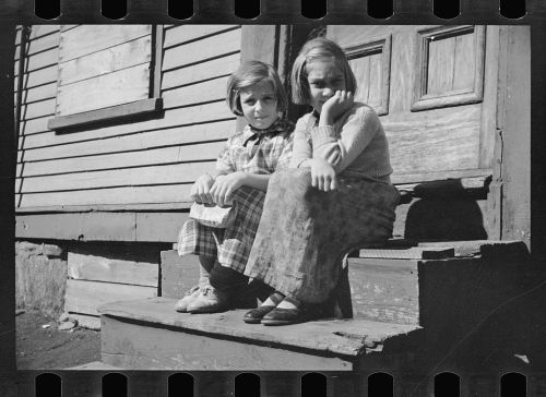 Two young girls sitting on wooden steps of a house, one holding a piece of paper, both looking at the camera.