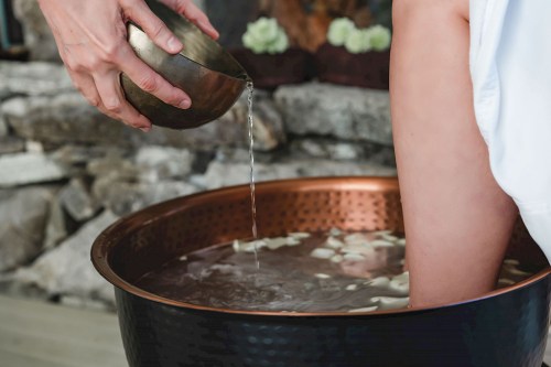 A person pours water from a bowl into a copper basin, with their foot submerged in the water.