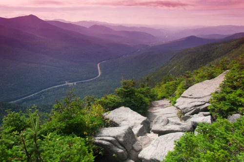 A scenic view of mountains and valleys at sunset, with rocky terrain and lush greenery in the foreground.