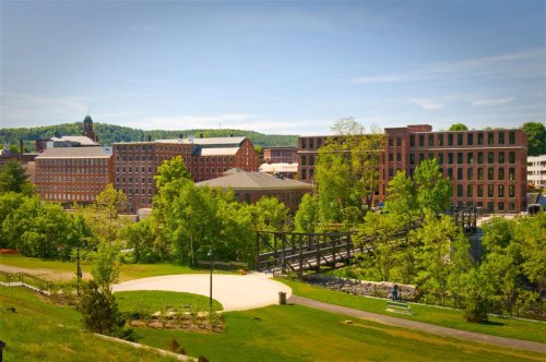 A scenic view of historic brick buildings surrounded by greenery and a bridge, under a clear blue sky.