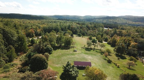 Aerial view of a lush green landscape with trees, a field, and a small pavilion under a blue sky with scattered clouds.