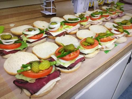A row of sandwiches on a countertop, layered with meats, cheese, lettuce, tomatoes, onions, and pickles.