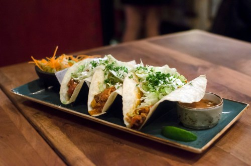 Three tacos filled with meat and topped with lettuce, served with a side of salad and a small bowl of sauce.