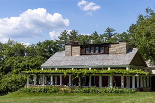 A charming house with a porch covered in greenery, surrounded by trees and a blue sky with fluffy clouds.