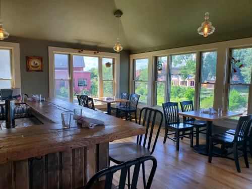 Cozy restaurant interior with wooden bar, tables, and large windows overlooking greenery and buildings outside.