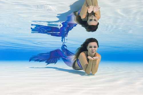 A mermaid with a purple tail blows a kiss while swimming underwater, reflected in the clear blue water.