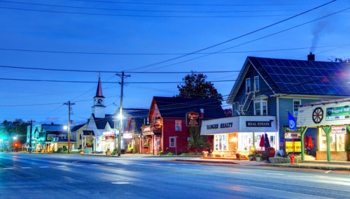 A quiet street at dusk featuring charming shops, a church with a steeple, and a house with solar panels.