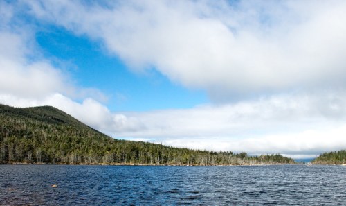 A serene lake surrounded by lush green forests and rolling hills under a partly cloudy sky.