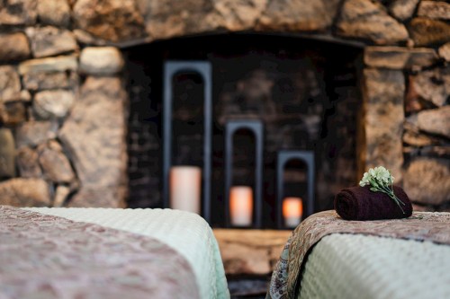 A serene spa setting with two massage tables, a stone fireplace, and soft candlelight in the background.