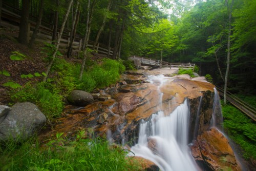 A serene waterfall cascades over smooth rocks, surrounded by lush green trees and a wooden fence in the background.