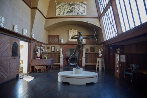 A spacious gallery featuring a central statue, sculptures on walls, and large windows allowing natural light.