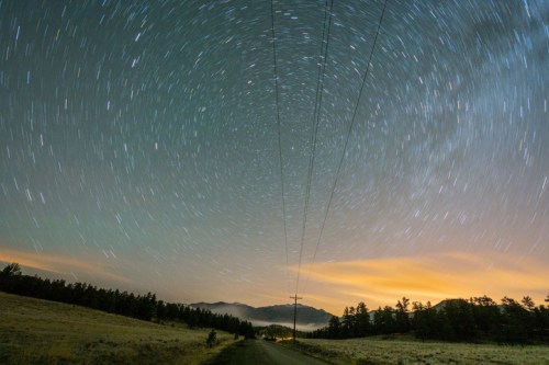 A long exposure shot of a starry night sky over a rural road, with power lines and mountains in the background.