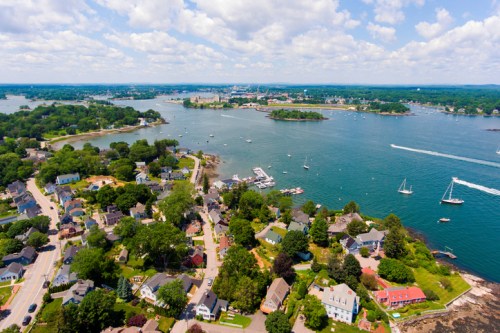 Aerial view of a coastal town with houses, boats, and lush greenery under a partly cloudy sky.