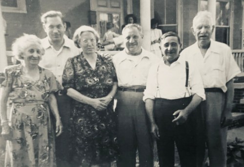 A black and white photo of six people standing together outdoors, smiling, in front of a house.