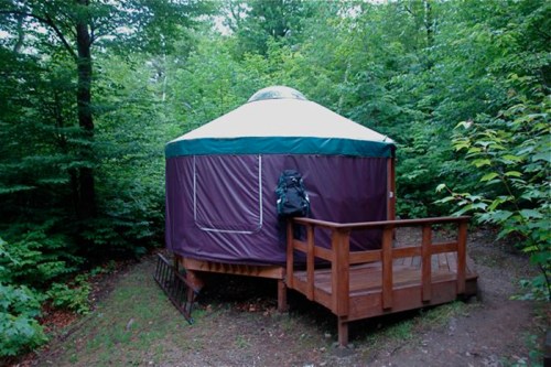A wooden yurt surrounded by lush green trees, featuring a small deck and a backpack resting against the wall.