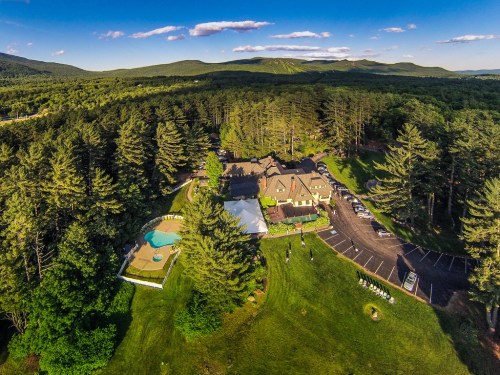 Aerial view of a lodge surrounded by trees, with a pool, parking area, and mountains in the background.